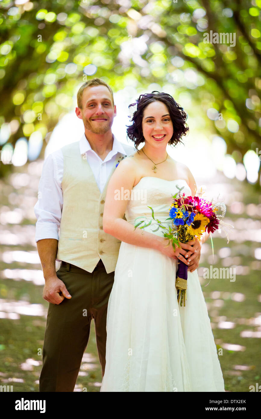 Braut und Bräutigam posieren für ein Porträt in einem hinterleuchteten Obstgarten am Tag ihrer Hochzeit in Oregon mit Licht, das durch die Bäume. Stockfoto