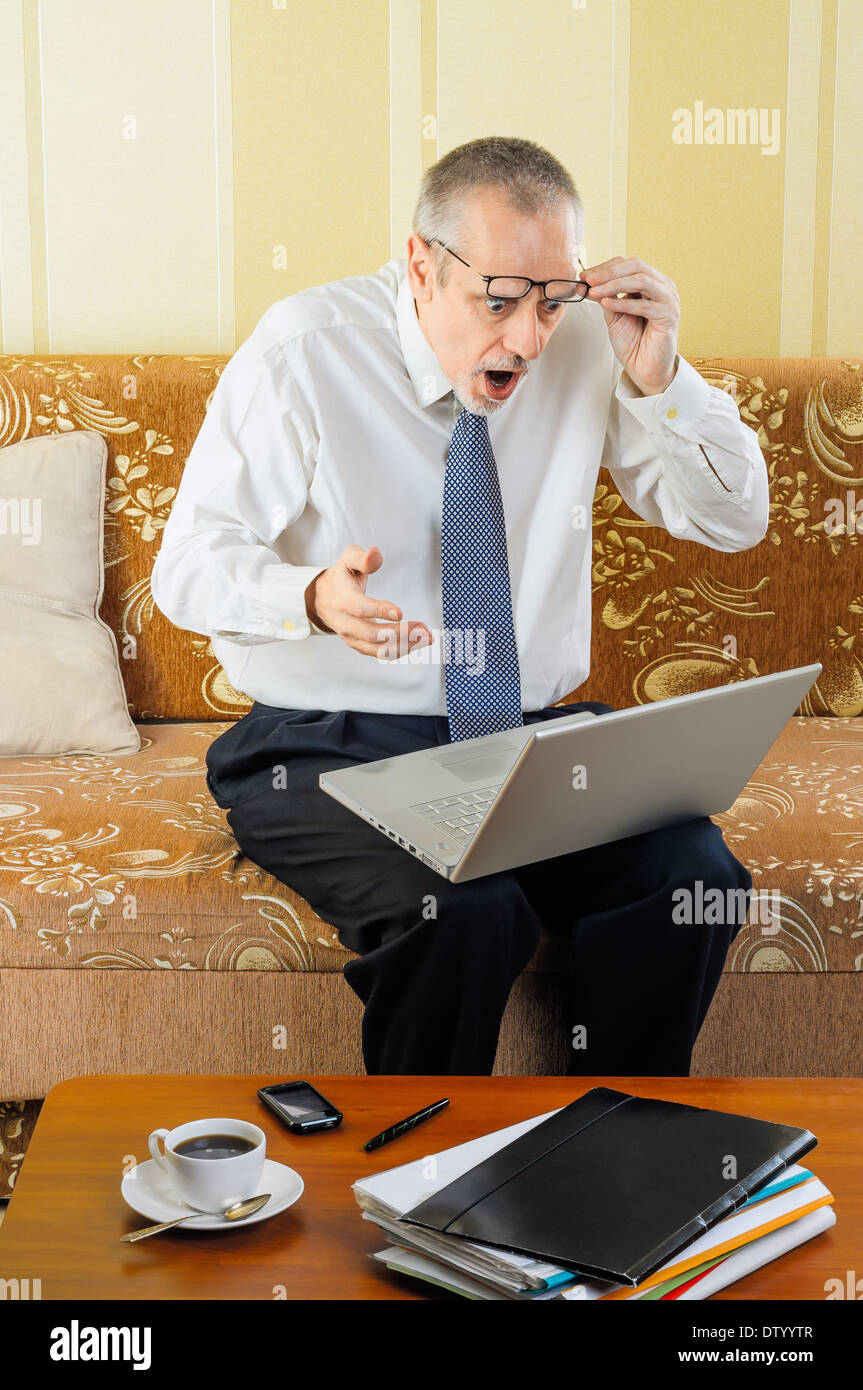 Ein hübscher senior Geschäftsmann arbeiten mit Computer zu Hause überrascht durch ein paar Neuigkeiten im Internet und Kaffee trinkfertig Stockfoto