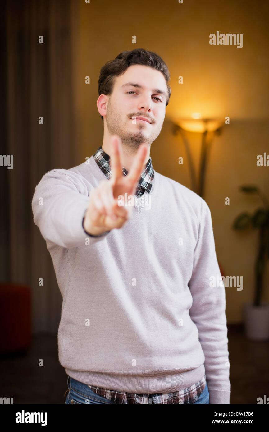 Attraktive junge Mann tut Frieden oder Sieg Schild mit zwei Fingern, indoor erschossen in einem Haus Stockfoto
