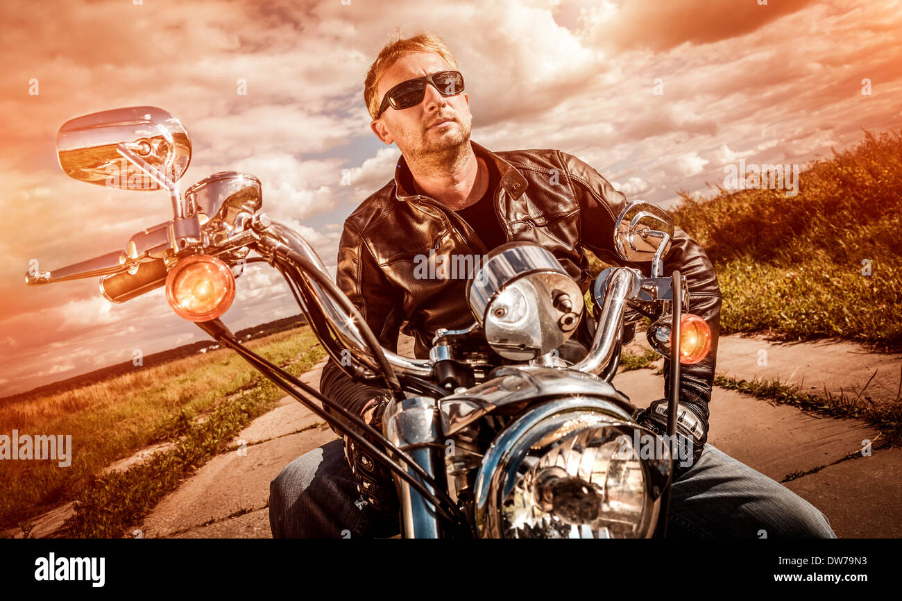 Biker-Mann trägt eine Lederjacke und eine Sonnenbrille sitzt auf seinem Motorrad, Blick auf den Sonnenuntergang. Stockfoto