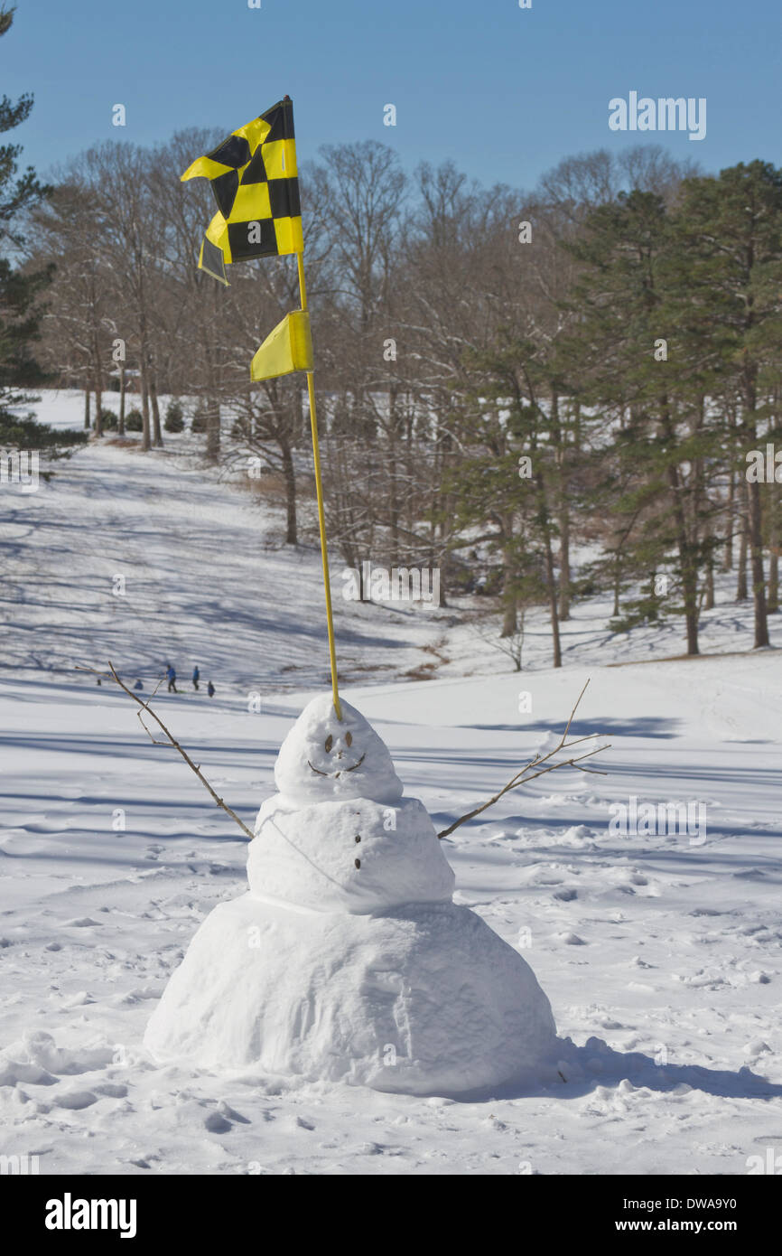 Ein Schneemann mit einem Golf Flagge sitzt lächelnd auf einem verschneiten Winter gold Kurs als Menschen Schlitten in der Ferne Stockfoto