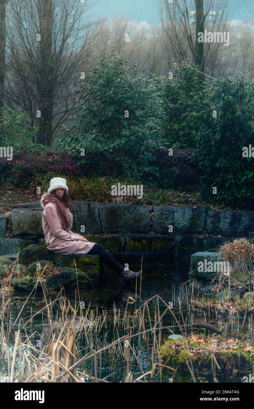 eine Frau in einem rosa Mantel sitzt an einem Teich Stockfoto