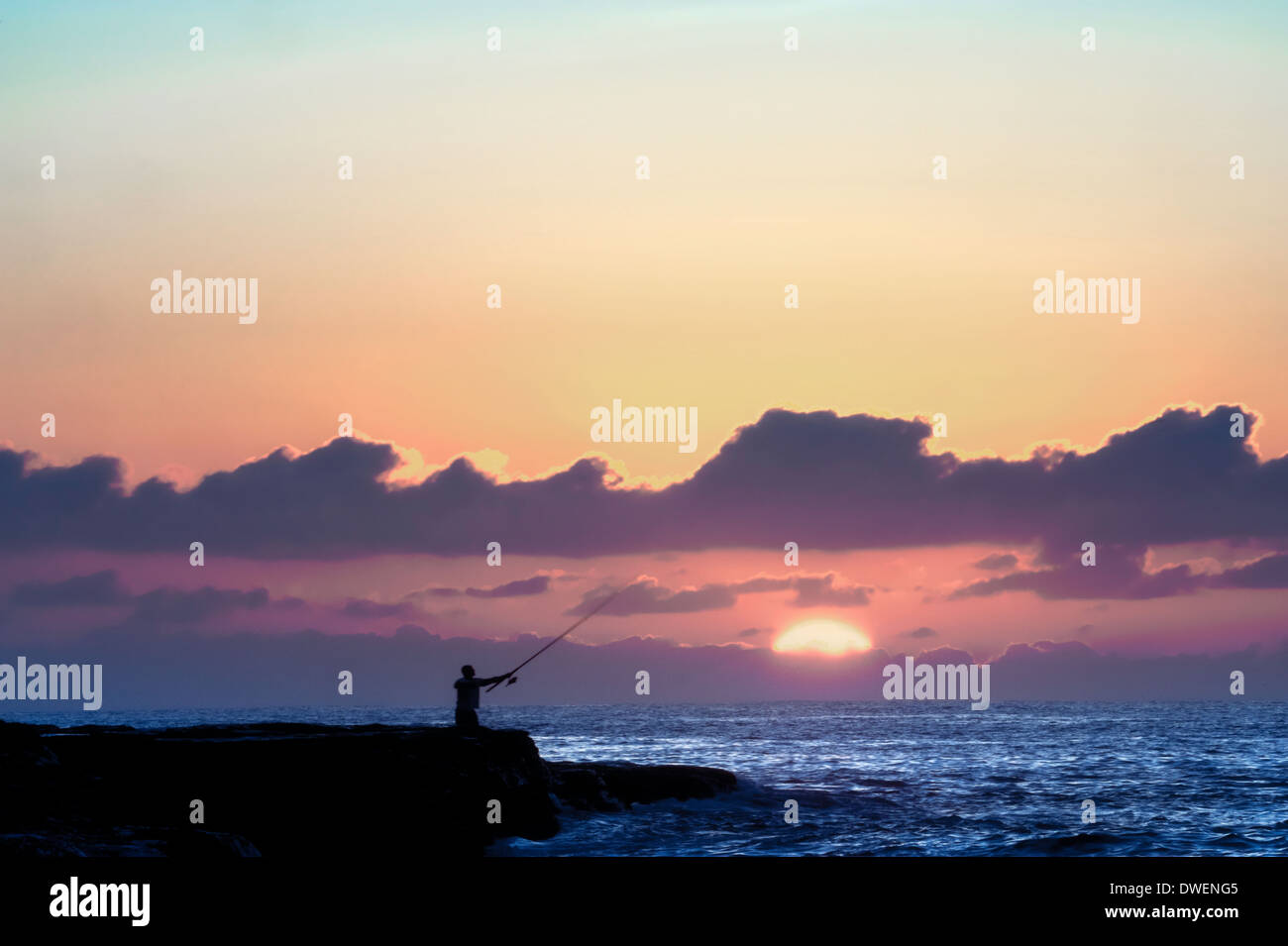 Ein einsamer Fels Fischer auswirft, wie die Sonne hinter Wolken am Horizont im Morgengrauen erscheint Stockfoto