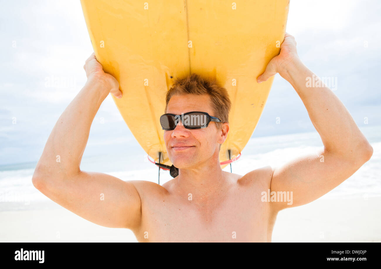 Männliche Surfer Surfbrett auf Kopf am Strand mit Meer und Himmel im Hintergrund ruht lächelnd Stockfoto
