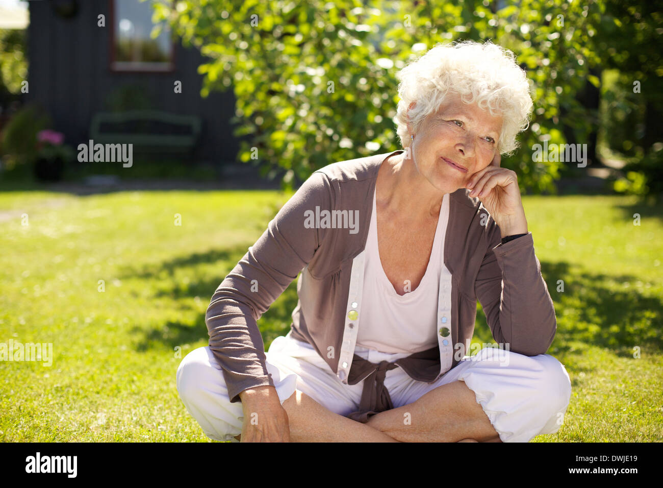 Ältere Frau sitzen im Freien auf dem Rasen wegschauen und denken. Ältere Frau Entspannung in Hinterhof Garten Tagträumen Stockfoto