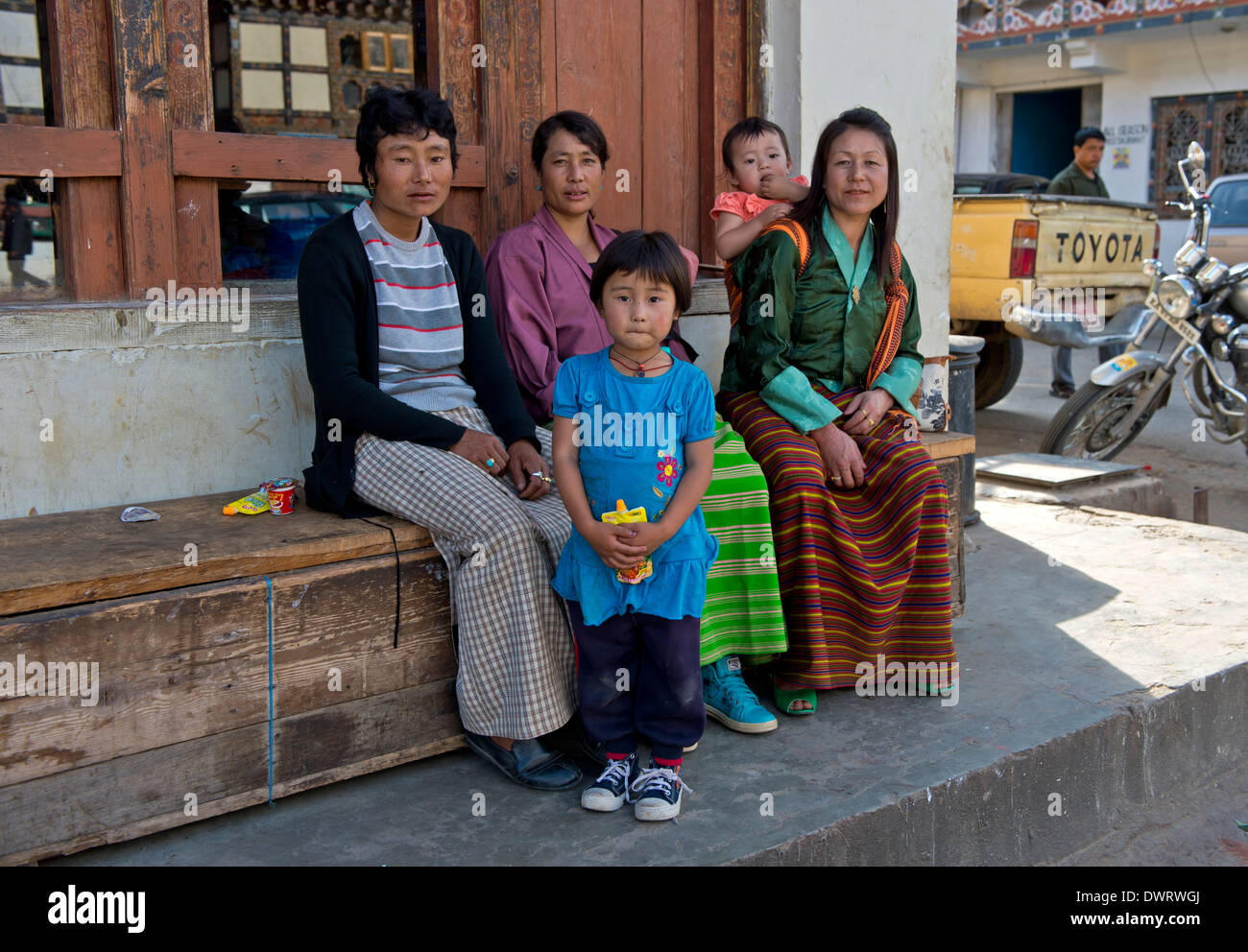 Mütter in Trachten mit Kindern sitzen auf einer Bank in der Hauptstraße von Paro, Bhutan Stockfoto