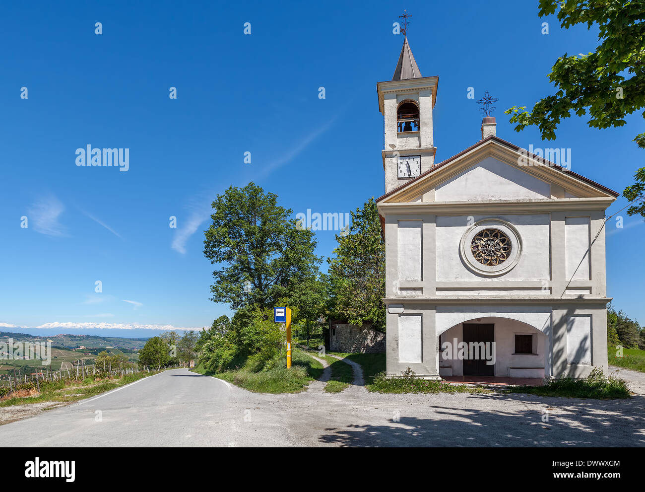Weiße Kapelle am Straßenrand und einspurige Straße unter blauem Himmel im Piemont, Norditalien. Stockfoto