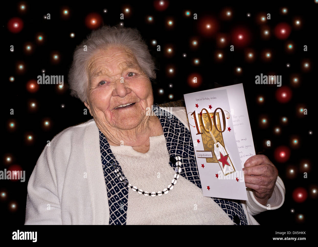 100 Jahre alt Happy Birthday Celebration lächelnd wachsam Dame mit einer '100' Glückwunschkarte & 100 farbige LED Party Lichter hinter Stockfoto
