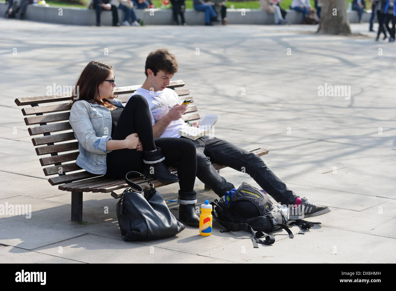 Ein junges Paar in Freizeitkleidung auf einer Bank sitzen. Der Mann SMS auf seinem Handy, während die Frau mit Blick auf das Telefon. Stockfoto