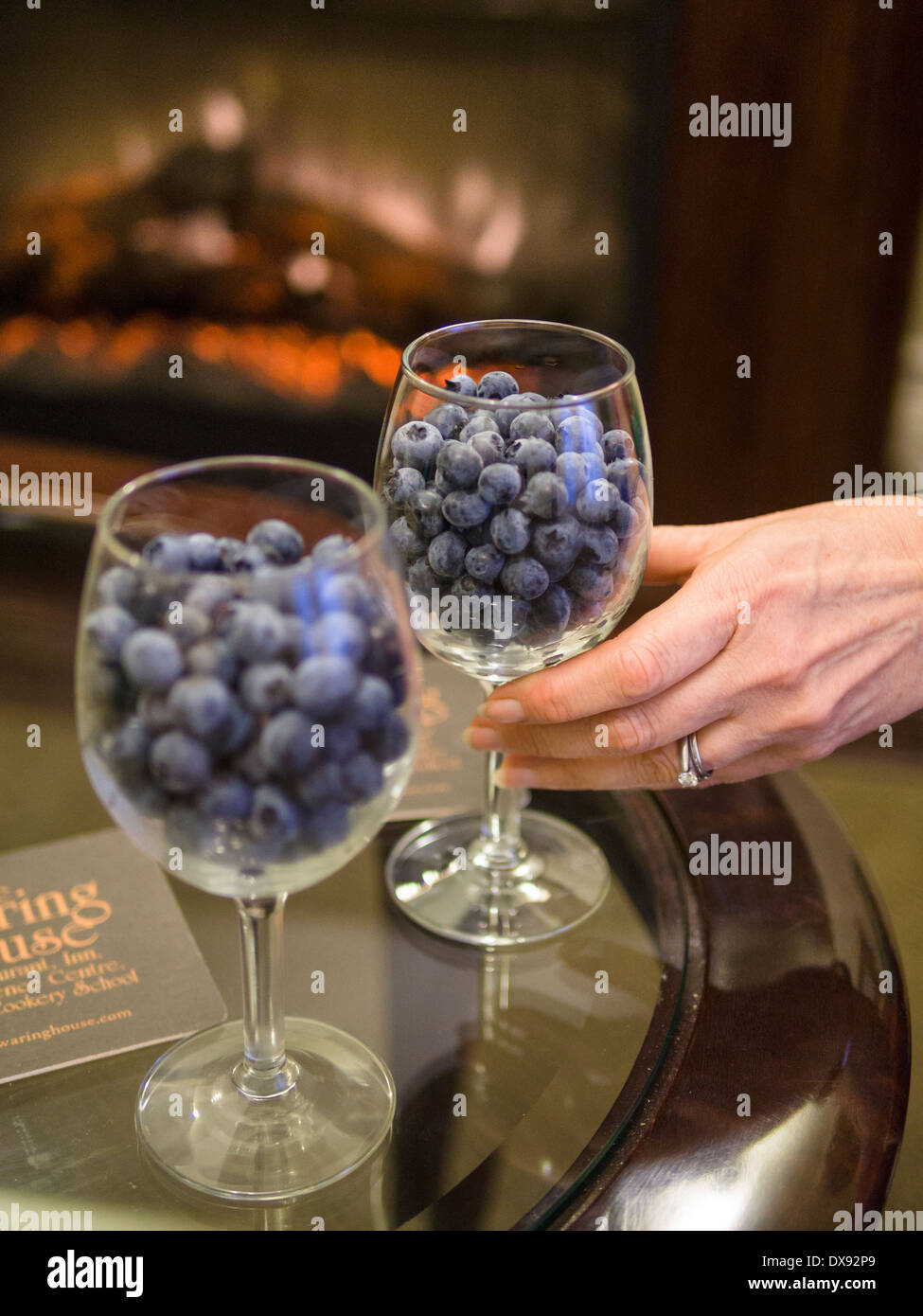 Romantische Heidelbeeren in Weingläsern. Heidelbeeren füllen Weingläser, die Hand einer Frau für einen von ihnen erreicht. Ein warmes Feuer glüht. Stockfoto