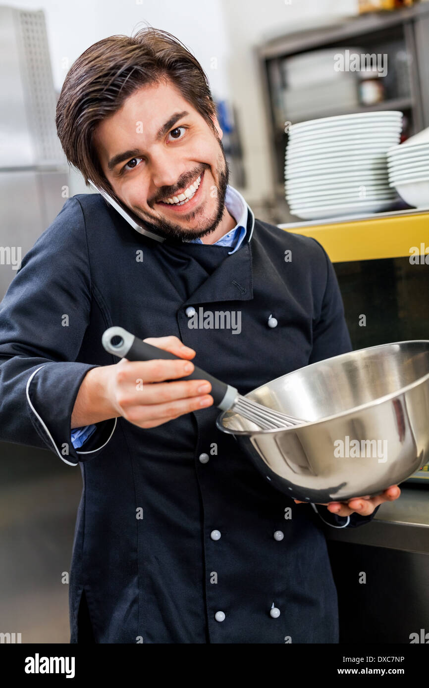 Professionelle Köchin oder Koch in einheitlichen stehend mit einem Edelstahl-Rührschüssel in der Hand, nehmen einen Anruf auf seinem Smartphone während der Vorbereitung Abendessen in einer Großküche in einem restaurant Stockfoto