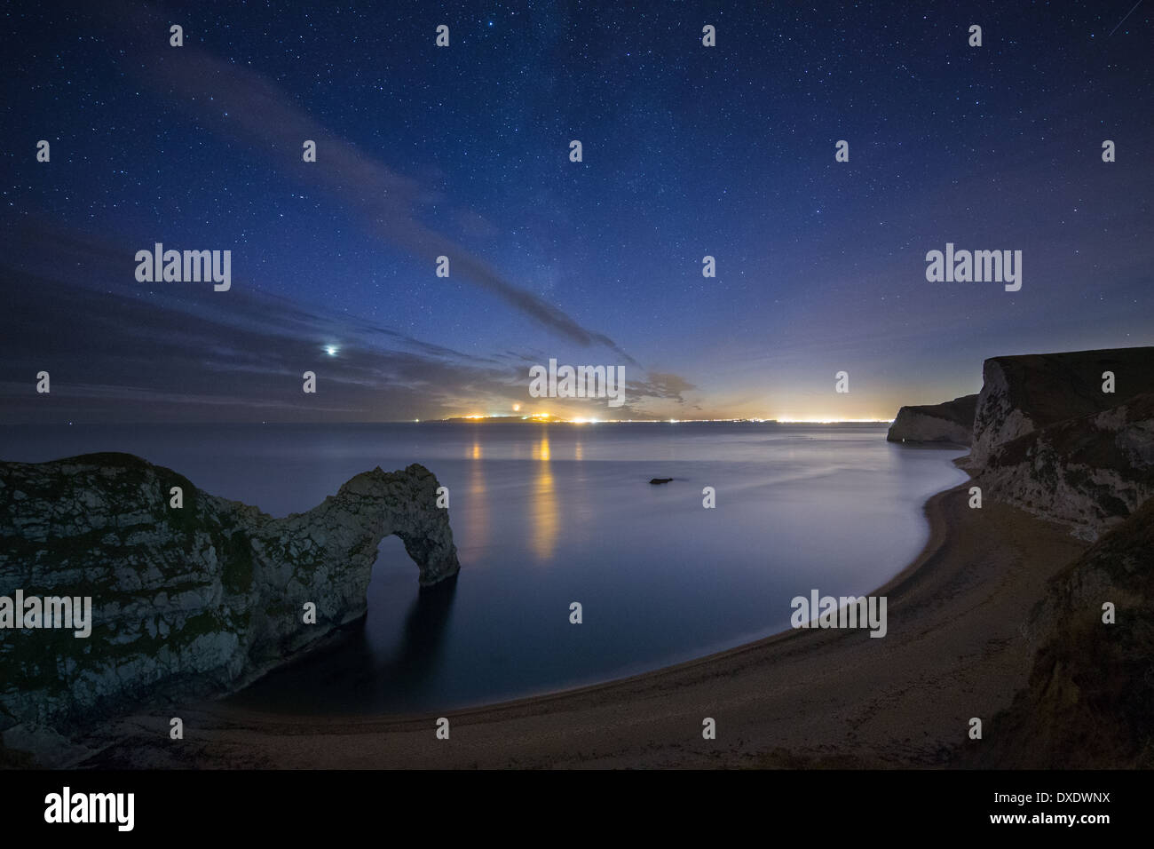 die Sterne und die Milchstraße über Durdle Door und die Jurassic Coast, mit den Lichtern von Weymouth & Portland darüber hinaus, Dorset, England Stockfoto