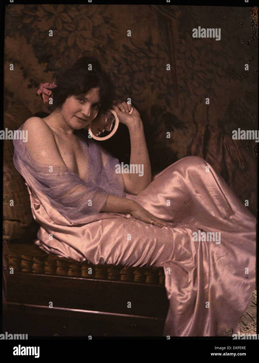 Dieses Vintage-Foto zeigt eine Frau in einem Satinkleid mit einem Spiegel und zeigt die Mode und den Stil der Vergangenheit. Das Bild spiegelt die Modetrends einer vergangenen Ära wider, die in einem historischen Druck festgehalten wurden. Stockfoto