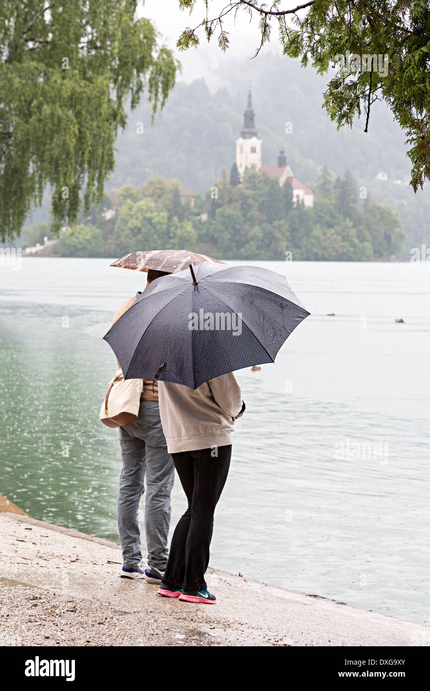 Zwei Menschen mit Regenschirmen in Regen am See mit der Kirche von Mary Pilgrimage, Bled, Slowenien Stockfoto