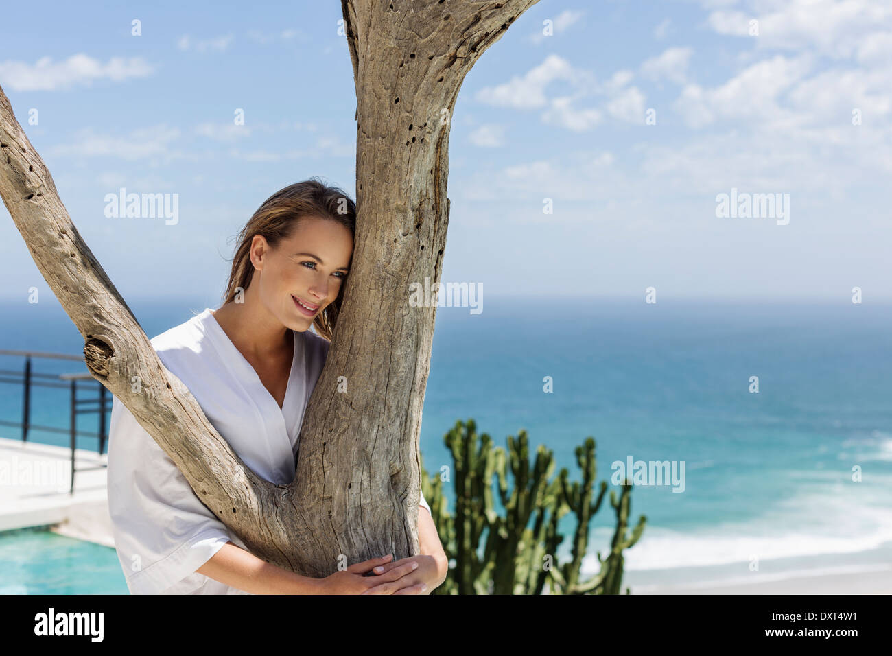 Lächelnde Frau in Bademantel umarmt Baum vor Ozean Stockfoto