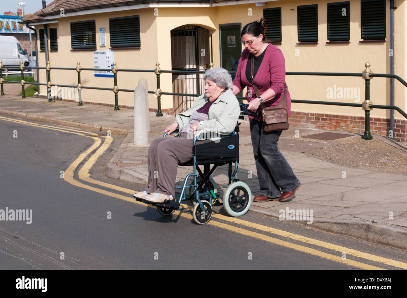 Eine ältere Frau in einem Rollstuhl mit Ihrem Betreuer oder Helfer überfahrt-Straße. Öffentliche behindertengerechte Toiletten im Hintergrund. Stockfoto