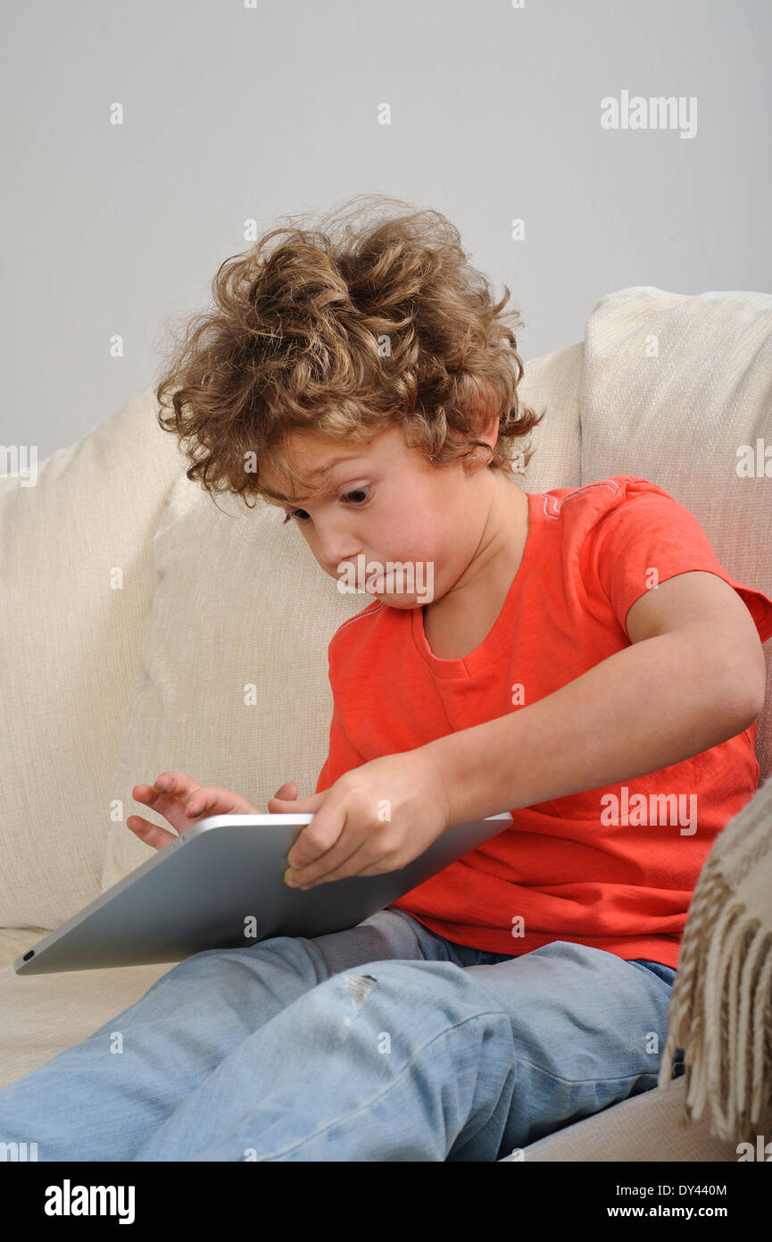 Eine ca. 8-9 Jahre alte Junge spielt Spiele auf einem Touch-Screen-Tablette. Er sitzt auf einem sofa Stockfoto