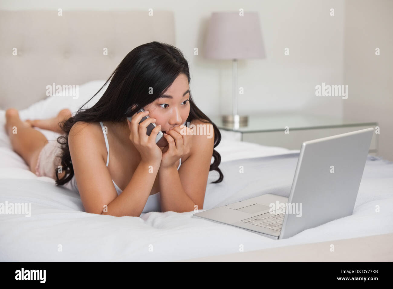 Ziemlich asian auf Bett telefonieren Blick auf laptop Stockfoto