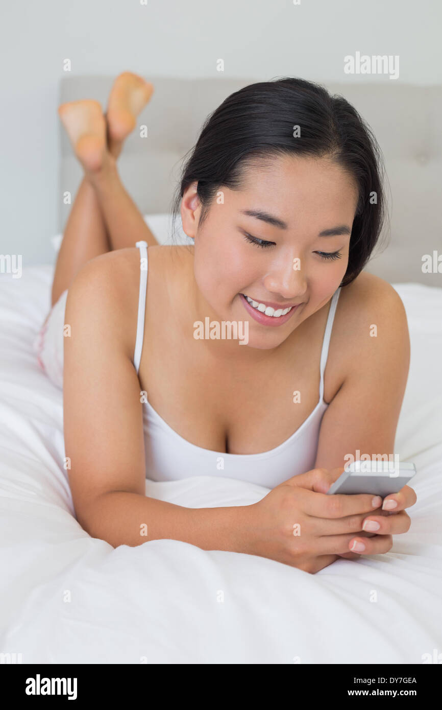 Lächelnde asiatische Frau liegend auf Bett SMS auf Handy Stockfoto