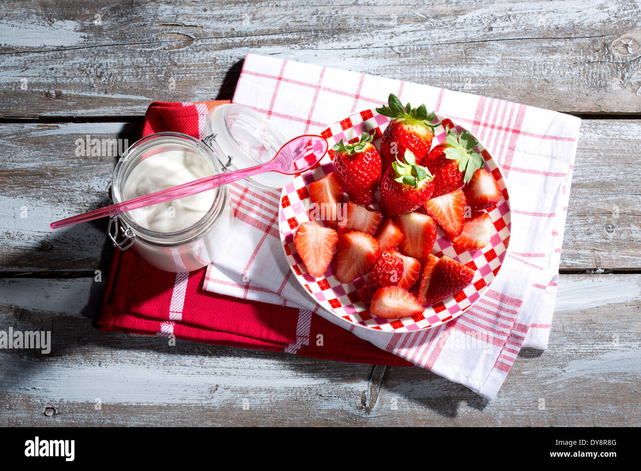 Glas Naturjoghurt mit Plastiklöffel, Teller mit in Scheiben geschnittenen und ganze Erdbeeren (Fragaria) auf Küchentücher und Holztisch Stockfoto