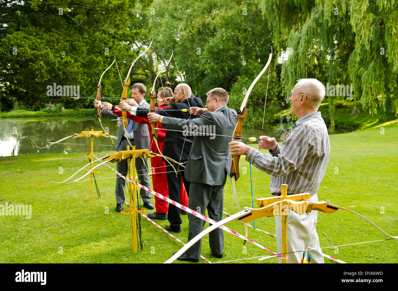 Gruppe von Männern lernen, Pfeil und Bogen schießen auf grünen Rasen vor Teich und Willow tree Stockfoto