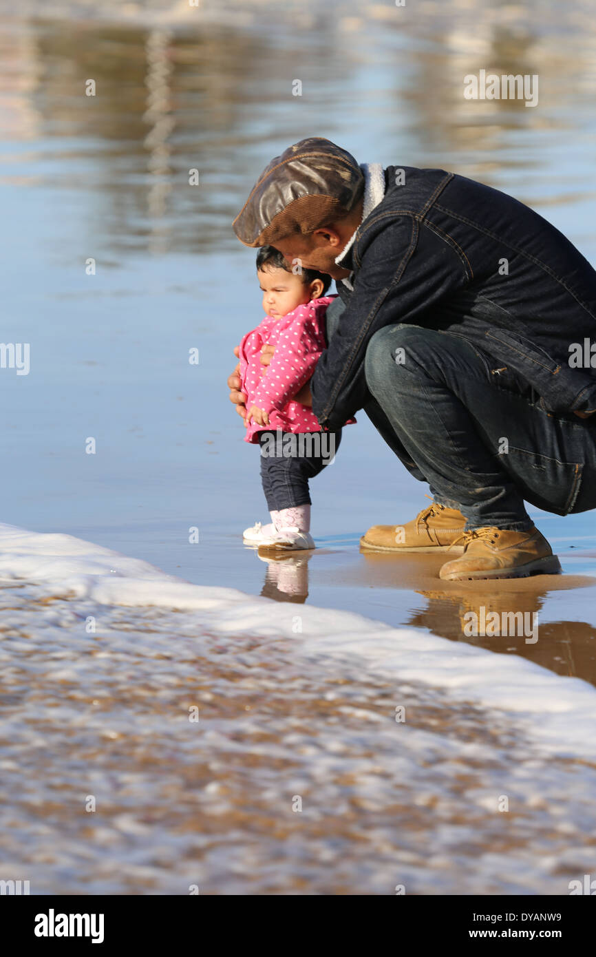 Vater Coying über Kleinkind Tochter auf einem nassen Sandstrand in Essaouira, Marokko, nicht zu sehen, eine Welle kommen bis zu allerletzten minu Stockfoto