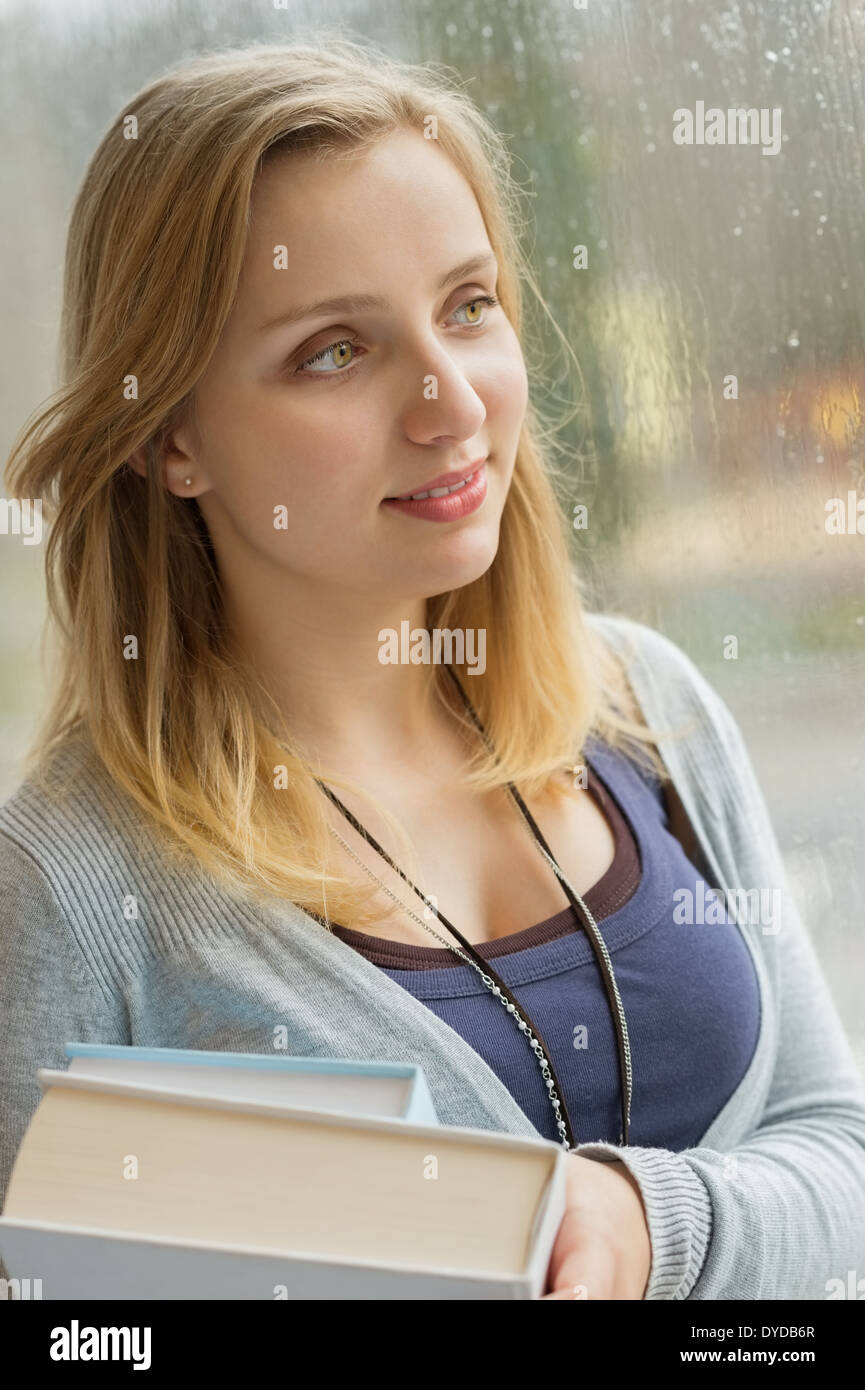 Nachdenkliche junge Studentin mit Büchern von Fenster während regnerischen Tag Stockfoto