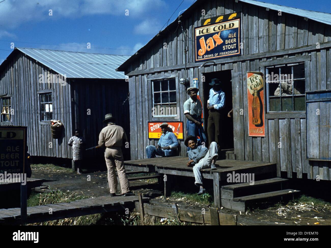 Wohnräume und "Juke Joint' für Wanderarbeiter, um während eines toten Saison sitzen; Belle Glade, Florida, USA. 1941. Foto zeigt Mann stehend mit Gewehr nach links. Zeichen werben Ice Cold Jax Ale Stout Bier und Eis kalte Coca Cola. Stockfoto
