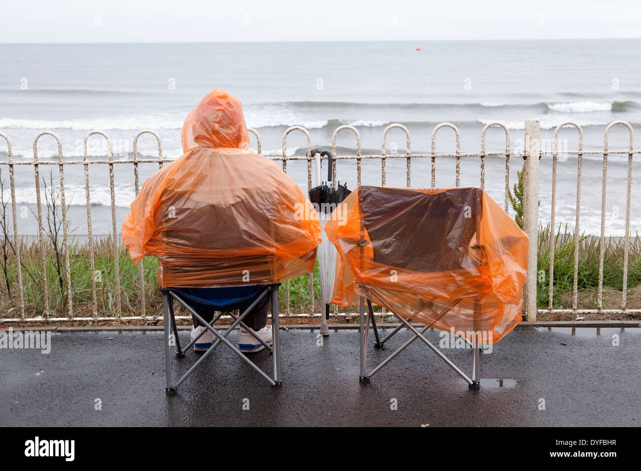 Eine Person sitzt in einem orange Wasserdicht am Meer im Regen. Großbritannien Stockfoto