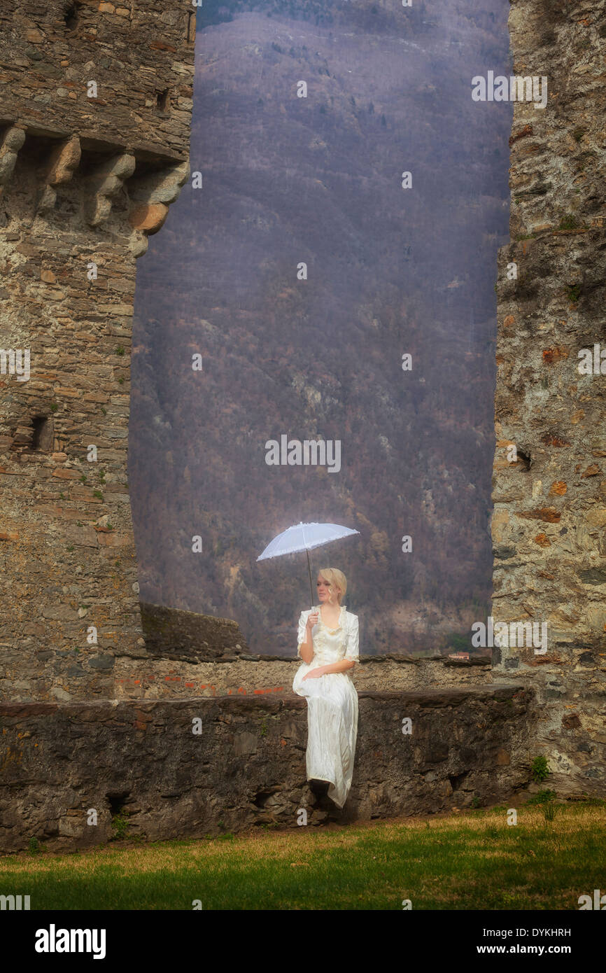 eine Frau in einem weißen Periode Kleid sitzt auf einer Mauer mit einem Sonnenschirm Stockfoto