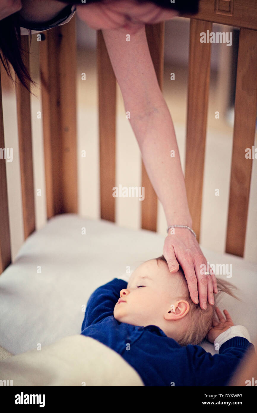 Baby schlafen im Kinderbett während Mutter beugt sich über und sanft streichelt ihren Kopf. Stockfoto