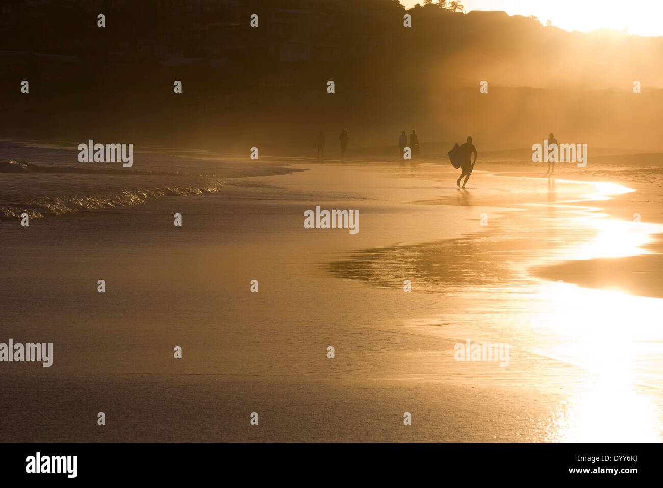 nebligen goldenen Sonnenuntergang auf Ebbe Strand Rand mit Lichtstrahlen Hintergrundbeleuchtung Surfer und Strandwanderer Mitte Abstand. Australien Stockfoto