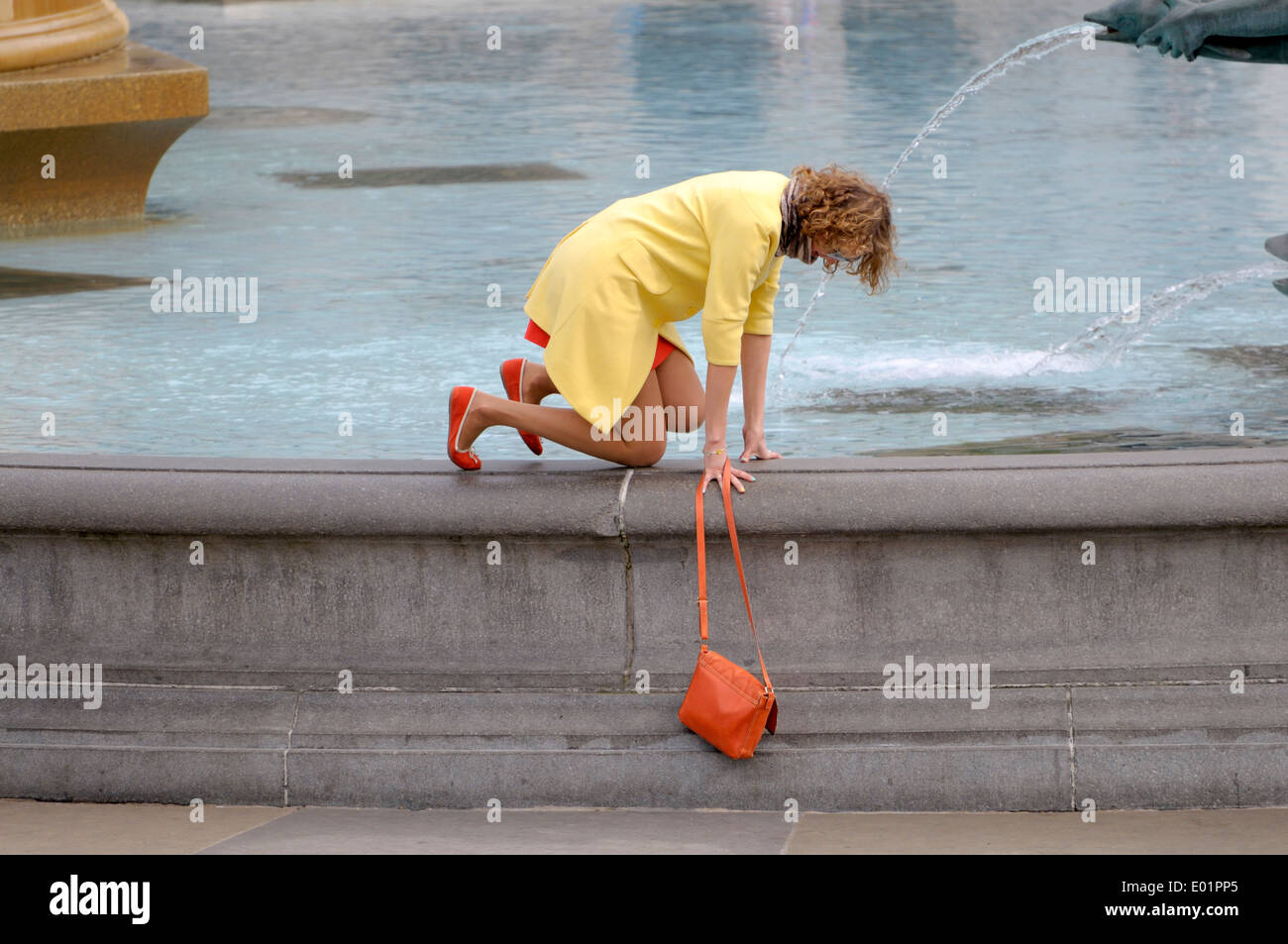 London, England, Vereinigtes Königreich. Trafalgar Square - Frau in einem gelben Mantel Klettern auf einen Brunnen (für ein Foto) Stockfoto