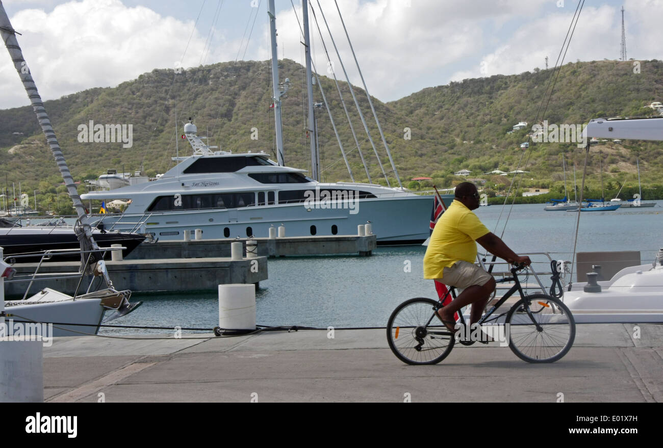 ein schwarzer Mann auf einem Fahrrad Zyklen vorbei an einer Luxus-Yacht im Hafen von Falmouth in Antigua Stockfoto