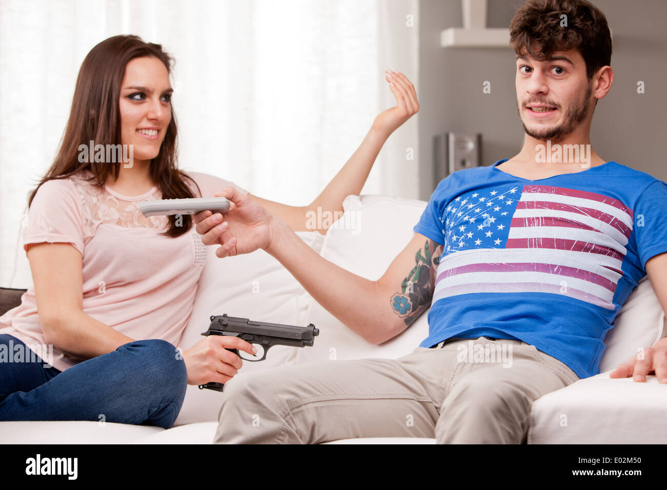 Frau auf einem Sofa anspruchsvoll sein Freund geben ihre Fernbedienung zwingt ihn mit einem Gewehr (lustige Szene) Stockfoto