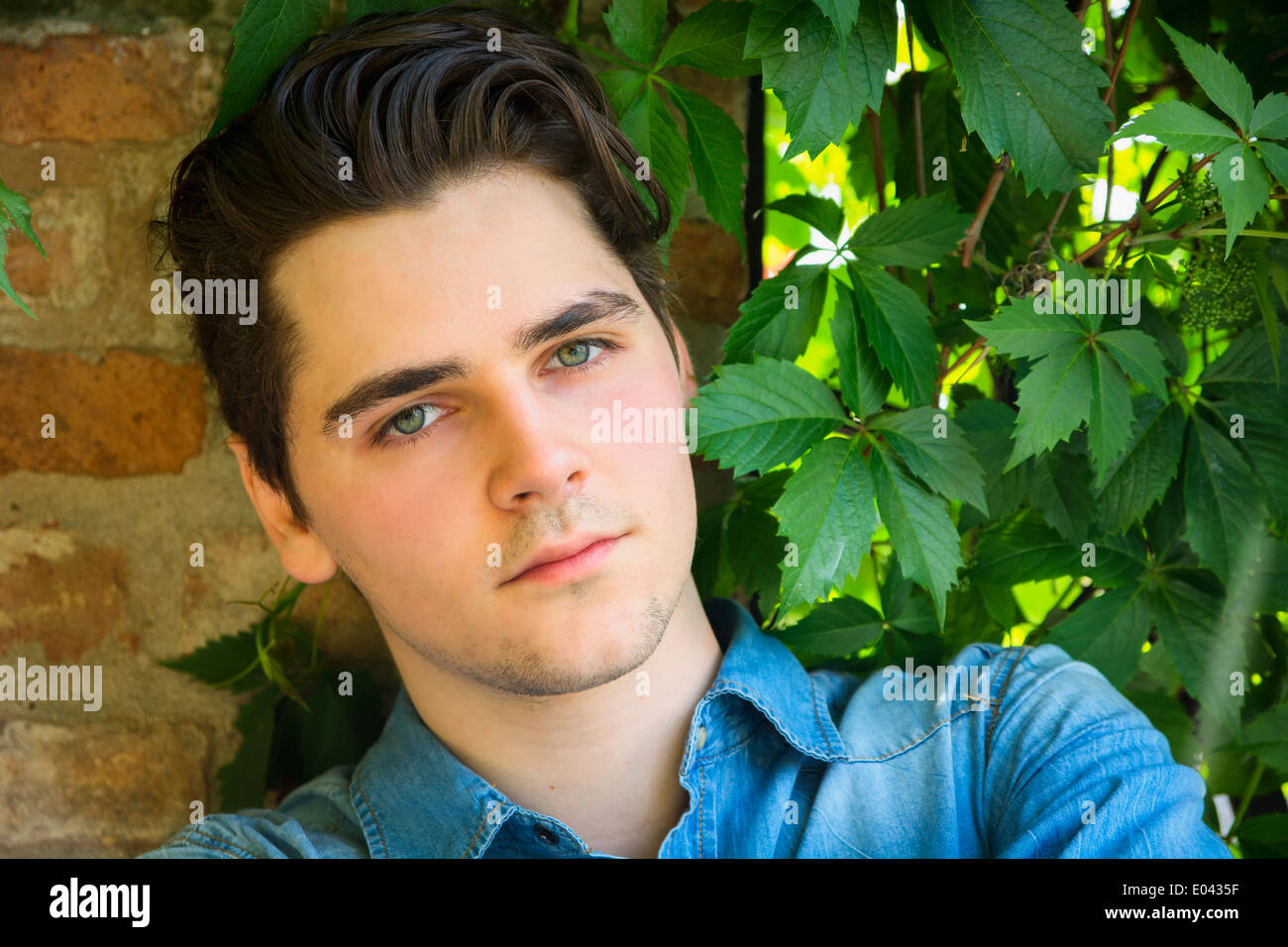 Attraktive junge Mann sitzt gegen die Mauer und Pflanze Blätter, Blick in die Kamera Stockfoto