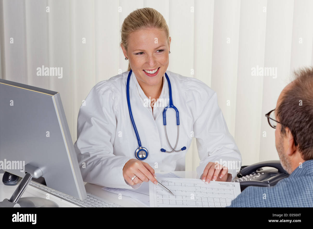 Ein junger Arzt mit Stethoskop in ihrer medizinischen Praxis. Im Gespräch mit einem Patienten, Eine Junge Aerztin Mit Stethoskop in Stockfoto