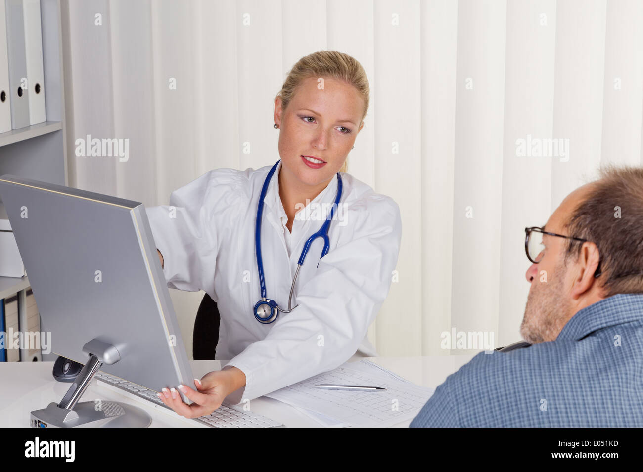 Ein junger Arzt mit Stethoskop in ihrer medizinischen Praxis. Im Gespräch mit einem Patienten, Eine Junge Aerztin Mit Stethoskop in Stockfoto