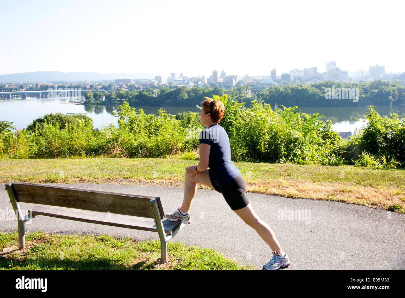 Weibliche Jogger erstreckt sich auf einer Bank vor der Ausführung auf einem Weg mit Blick auf die Skyline einer Riverview Stadt an einem dunstigen, heißen Sommertag. Stockfoto