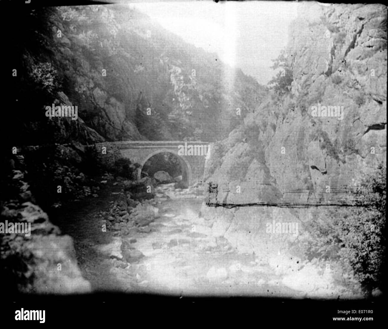 Ein Fluss fließt durch eine enge Schlucht zwischen zwei felsigen Hängen, über die eine Steinbrücke überquert. Diese malerische Szene, die in der Region Ariège zu finden ist, ist in der Bibliothèque de Toulouse erhalten und bietet einen Einblick in die natürliche Schönheit und historische Architektur der Region. Stockfoto
