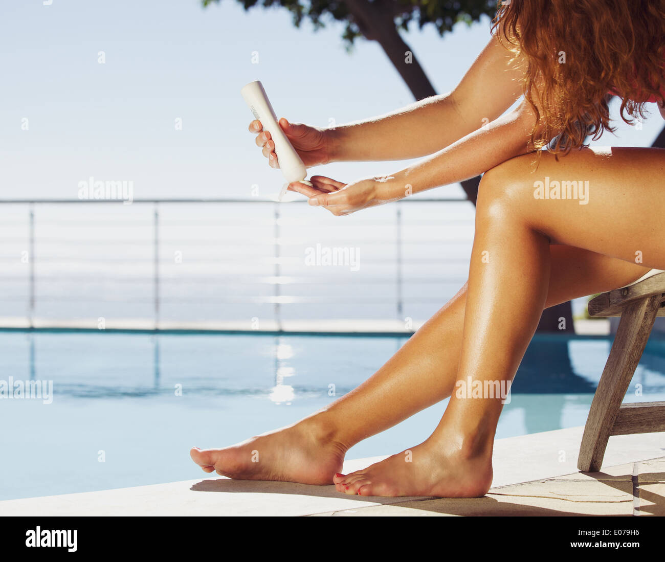 Frau sitzt auf Liegestuhl durch das Schwimmen pool Anwendung Sonnencreme auf ihre Beine. Weibliches Model Sonnenbaden am Pool. Stockfoto