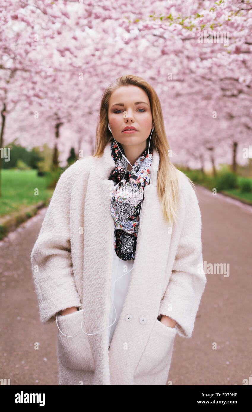 Hübsche junge weibliche Modell tragen lange Mantel Musikhören gleichzeitig blühen Frühlingsgarten. Stockfoto