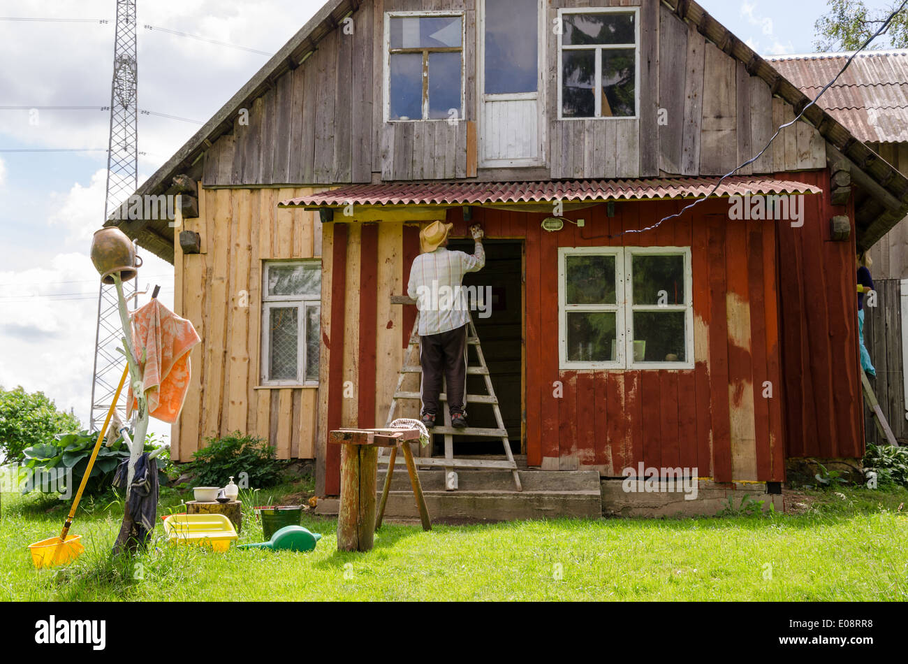 Maler Bauer Mann mit Hut auf Leiter Farbe alten hölzernen ländlichen Gehöft Hauswand mit Pinsel Pinsel. Stockfoto