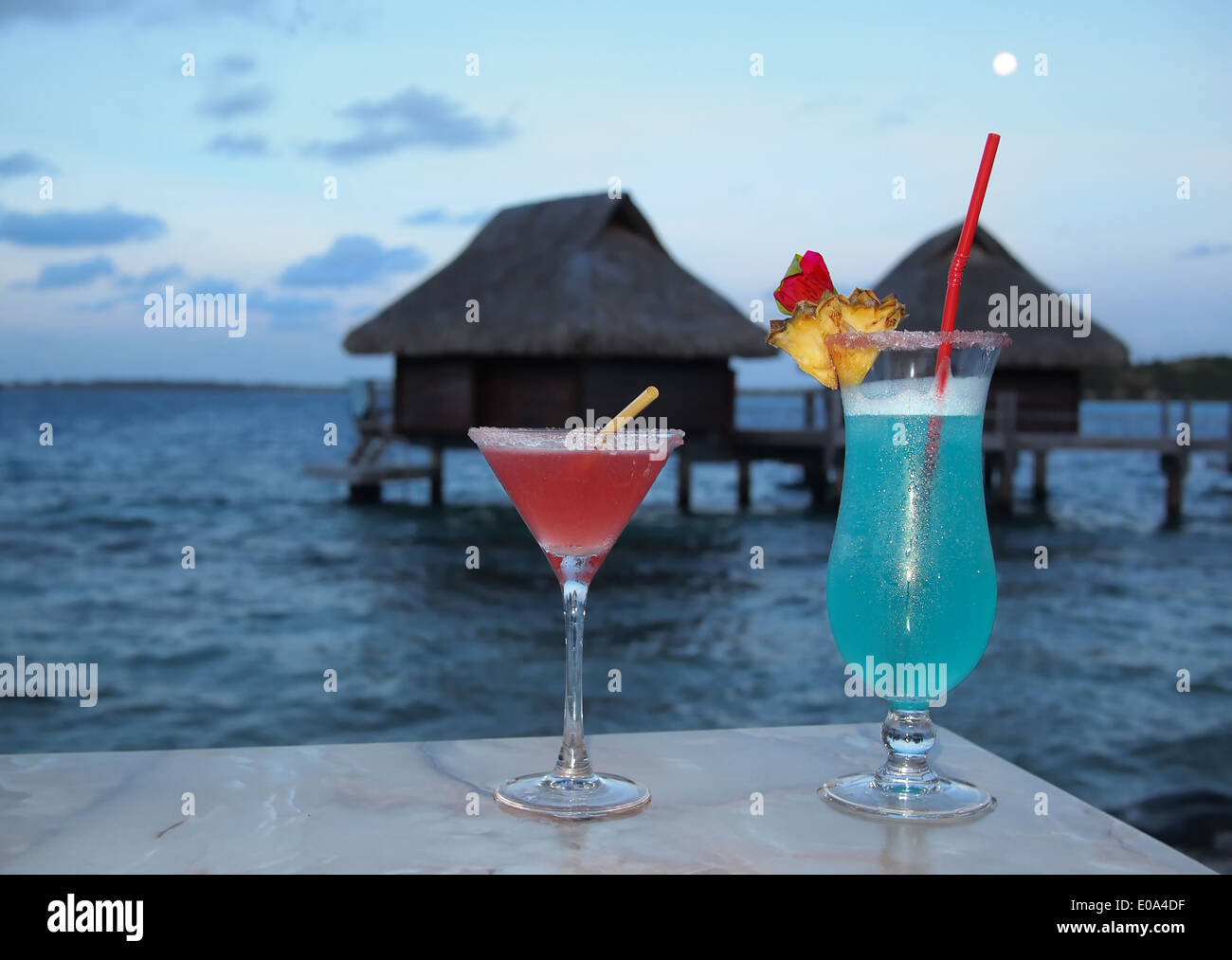 Flitterwochen. Zwei Cocktails auf einem Tisch in der Abenddämmerung mit im Hintergrund strohgedeckten Wasserbungalows auf einer tropischen Insel. Stockfoto