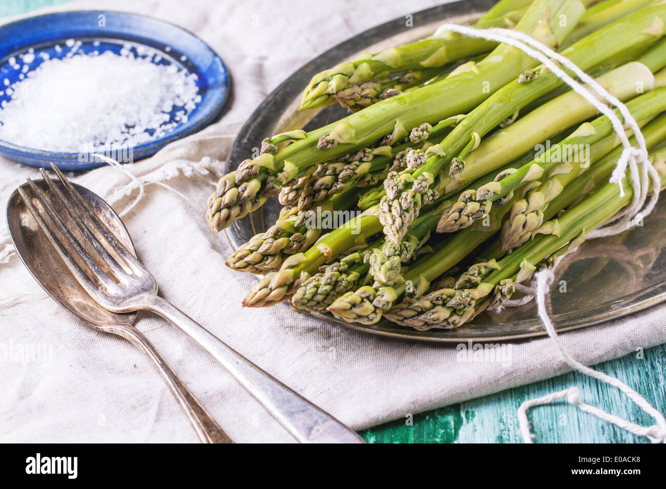 Haufen junger Grüner Spargel auf Vintage Tablett serviert mit Keramikplatte von Meer Salz und Silber Besteck über grüne Holztisch. Stockfoto