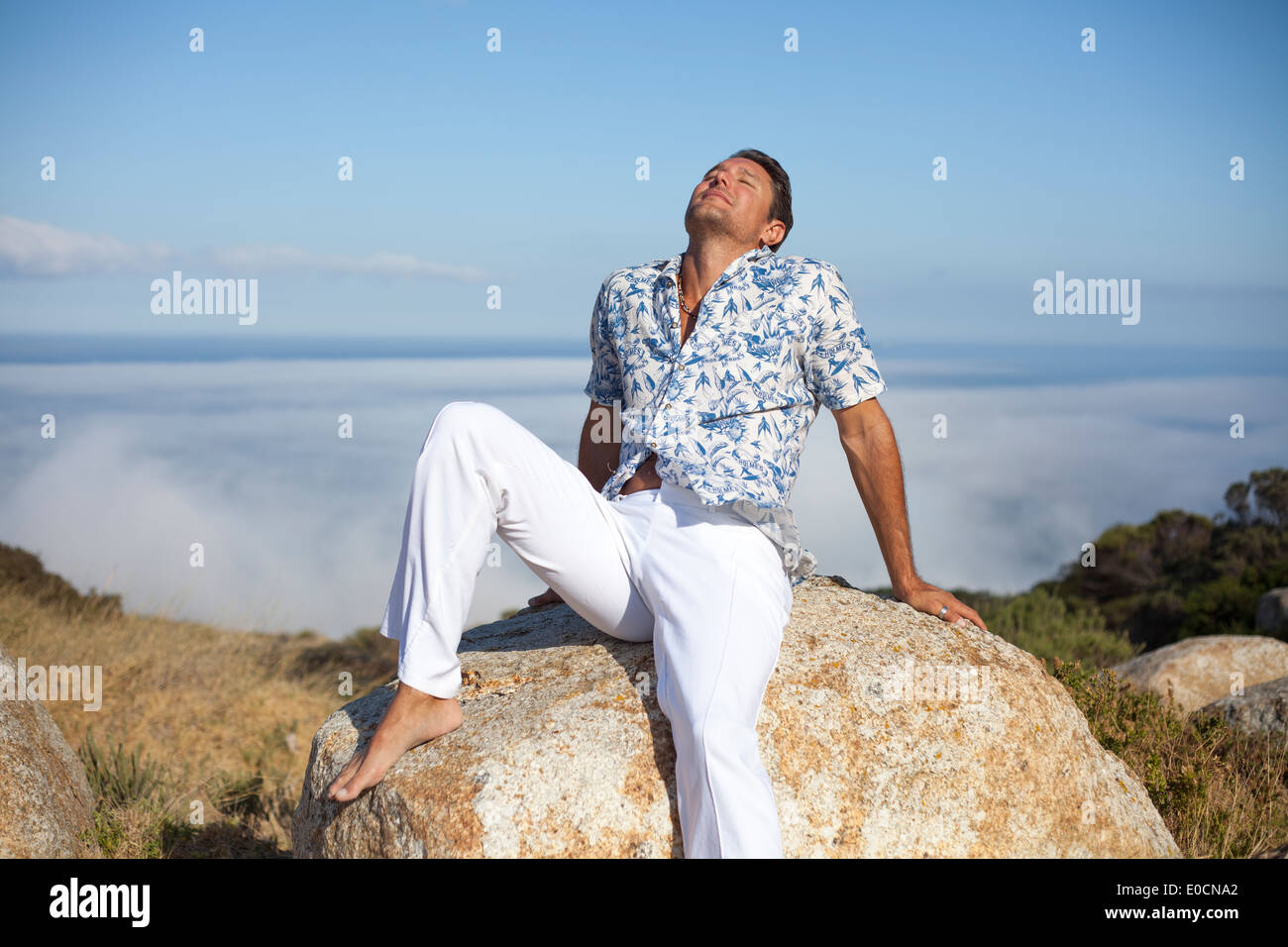 Mann sitzt auf einem Felsen oberhalb der Nebelgrenze Rollen über das Meer, auf einem Berg Stockfoto