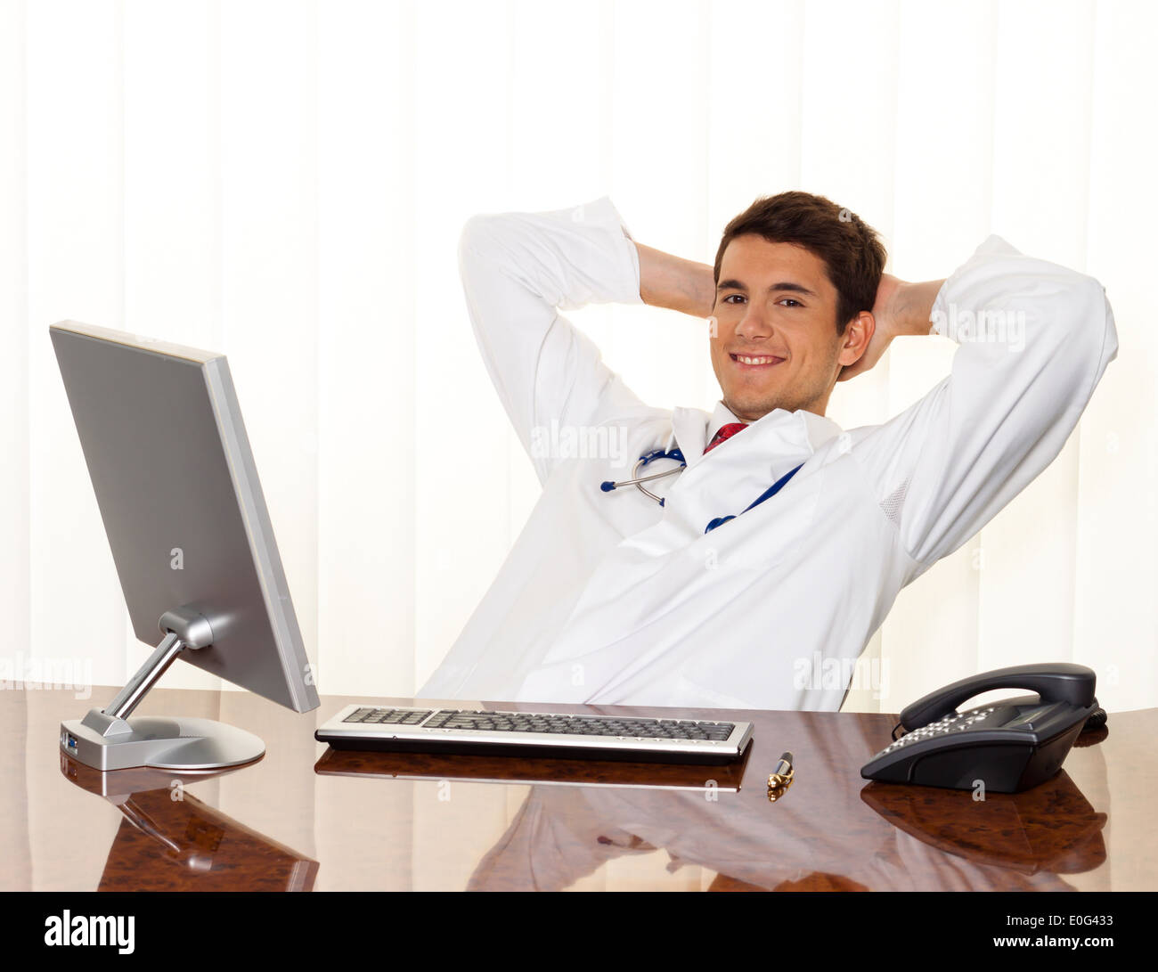 Ein erfolgreicher junger Arzt sitzt mit Schreibtisch und einem Lächeln, Ein Erfolgreicher Junger Arzt Sitzt Bei Schreibtisch Und Laechelt. Stockfoto