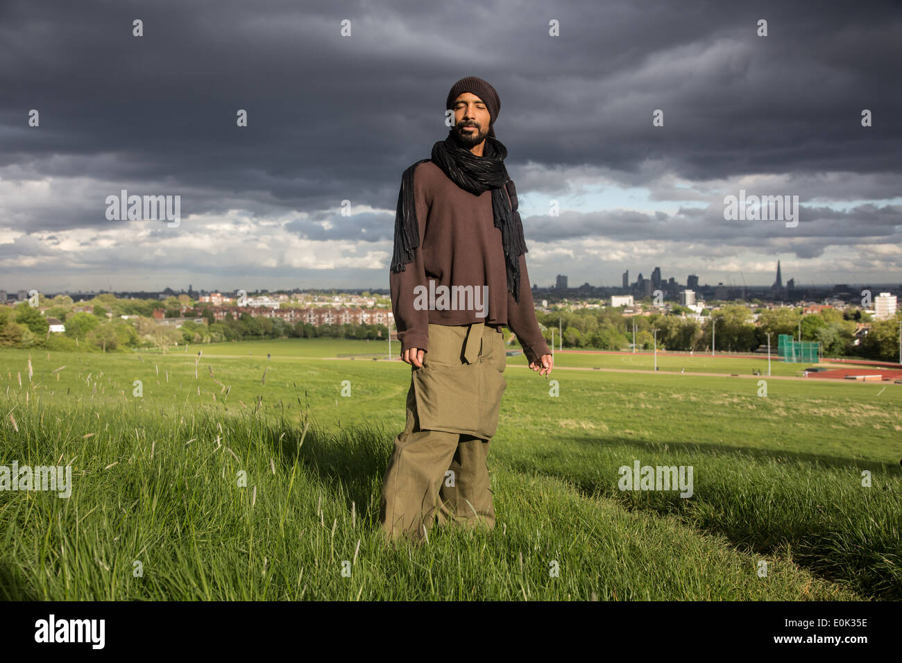 Gemischte Rassen Mann auf Hampstead Heath mit Augen geschlossen tragen Kopftuch und sackartige Kleidung mit stimmungsvollen Wolken im Himmel hinter Stockfoto