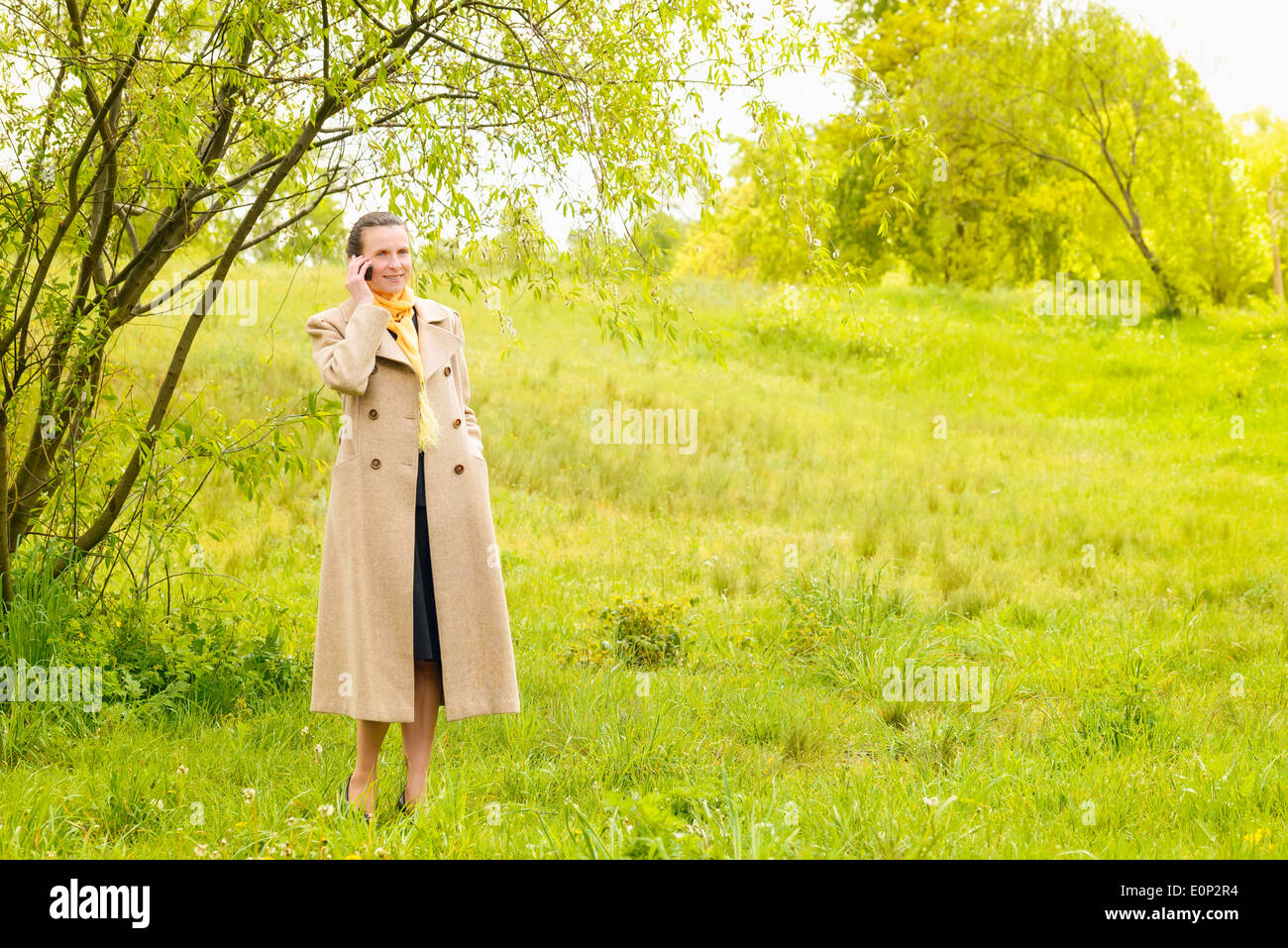 Eine elegante senior Geschäftsfrau, mit einem Mantel und einem gelben Schal, telefonieren mit ihrem Handy im Park unter der Frühlingssonne Stockfoto