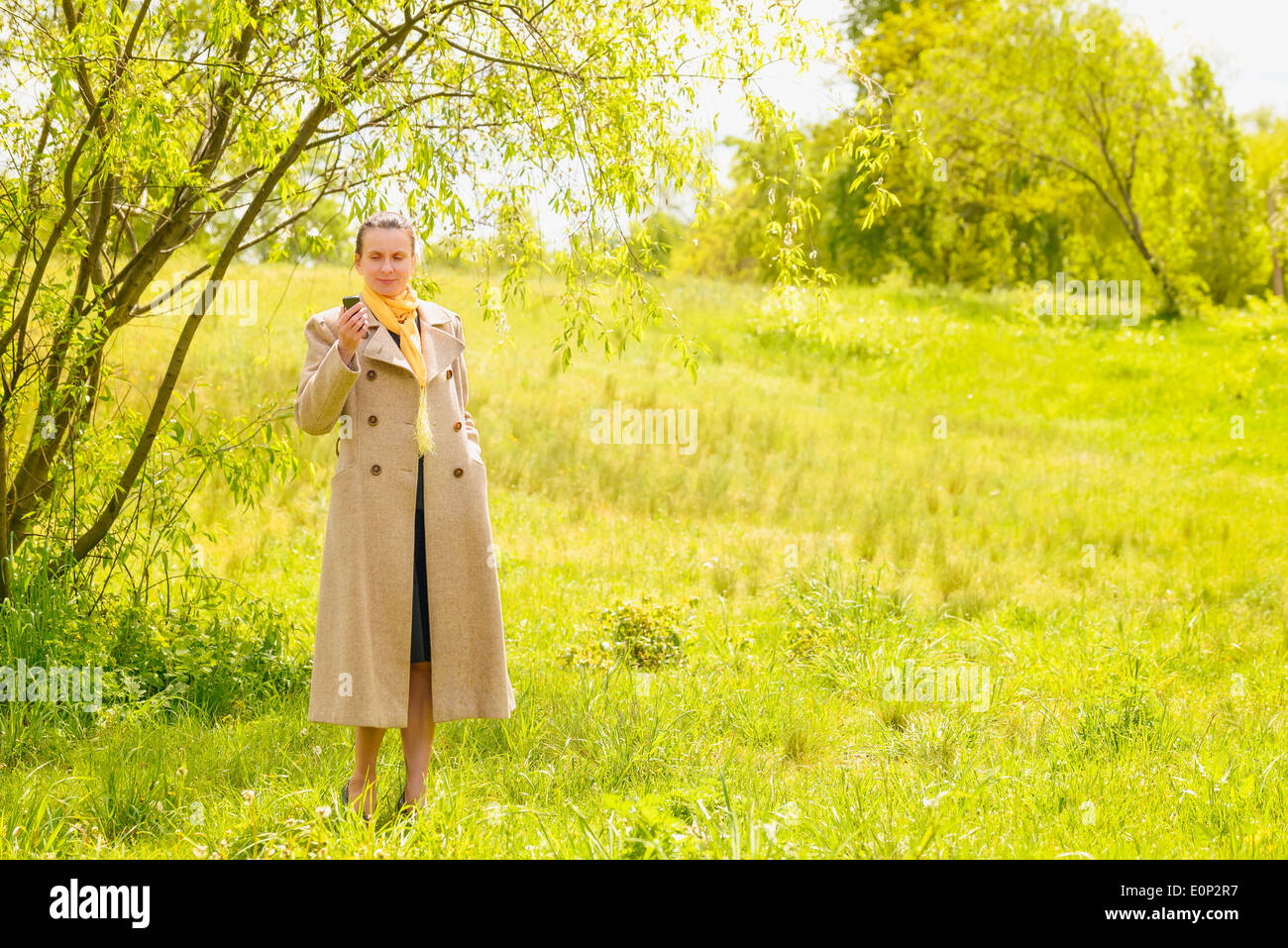 Eine elegante senior Geschäftsfrau, mit einem Mantel und einem gelben Schal, Blick auf ihr Handy im Park unter der Frühlingssonne Stockfoto